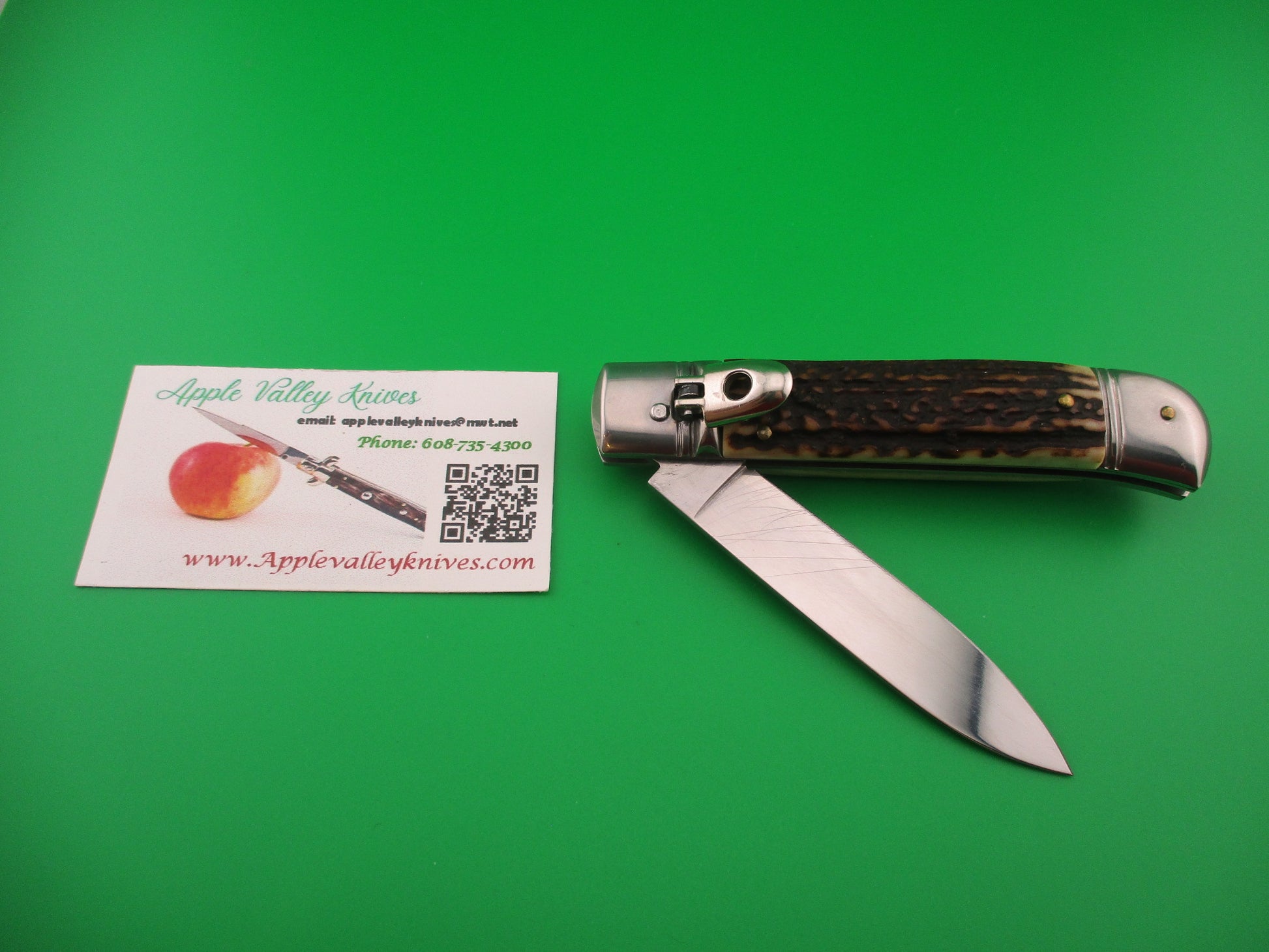 Foldable knife with staghorn handle on a green background, next to an Apple Valley Knives business card.