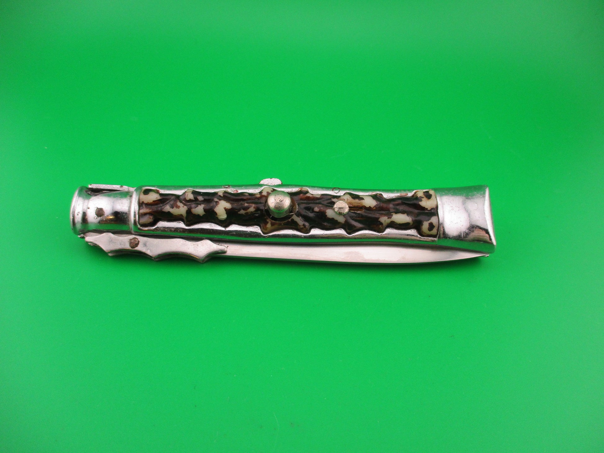 A folding pocket knife with a polished silver-toned metal body and a decorative handle featuring black, brown, and white marbled inlay, shown against a solid green background.